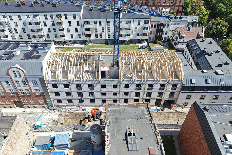 Zdjęcie z lotu ptaka przedstawia plac budowy nowoczesnego budynku wielorodzinnego z wyraźnie widoczną, drewnianą konstrukcją dachu. Nad inwestycją góruje niebieski żuraw wieżowy, a w otoczeniu obiektu znajdują się inne, gotowe już budynki mieszkalne oraz zaplecze budowlane.