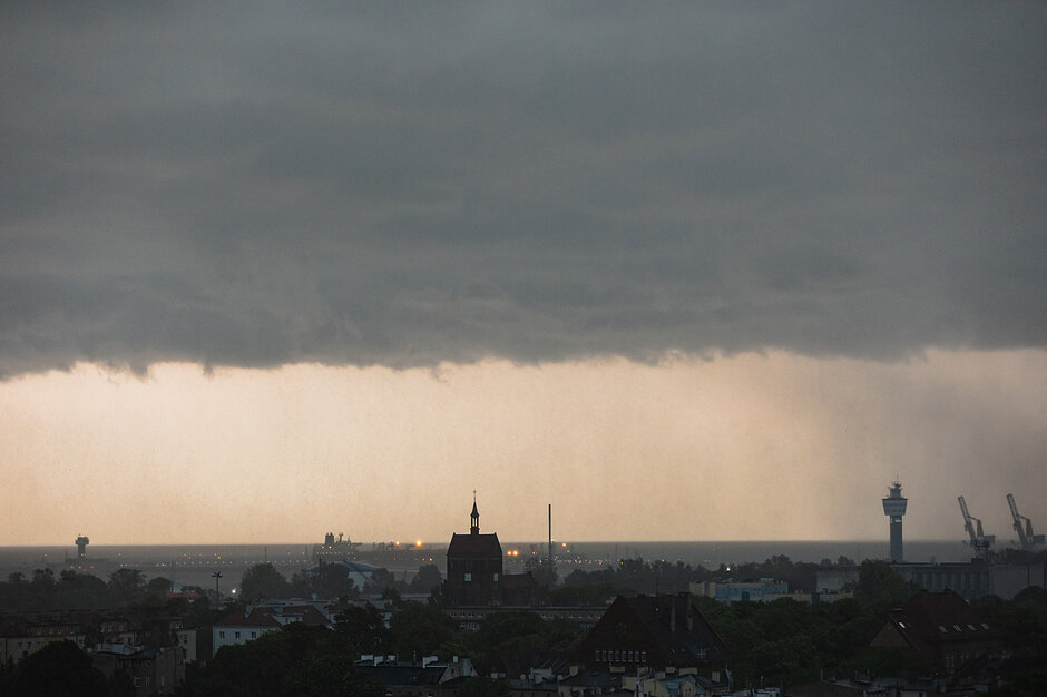 Zdjęcie przedstawia panoramę nadmorskiego miasta lub portu pod ciemnym, burzowym niebem, z którego w oddali widoczna jest ściana deszczu. Na pierwszym planie odznaczają się sylwetki budynków, w tym charakterystyczna wieża kościoła oraz portowe żurawie i wieża kontrolna.