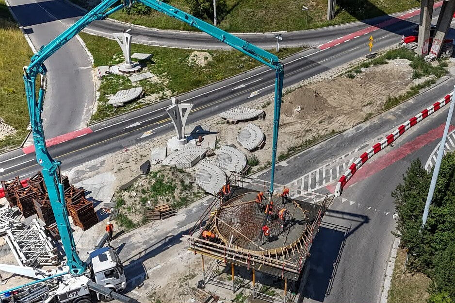 Zdjęcie przedstawia plac budowy infrastruktury drogowej, gdzie za pomocą pompy do betonu zalewana jest okrągła platforma zbrojeniowa. Wokół konstrukcji pracuje grupa robotników w odzieży ostrzegawczej, a w tle widoczne są gotowe elementy architektoniczne oraz sąsiadujące drogi.