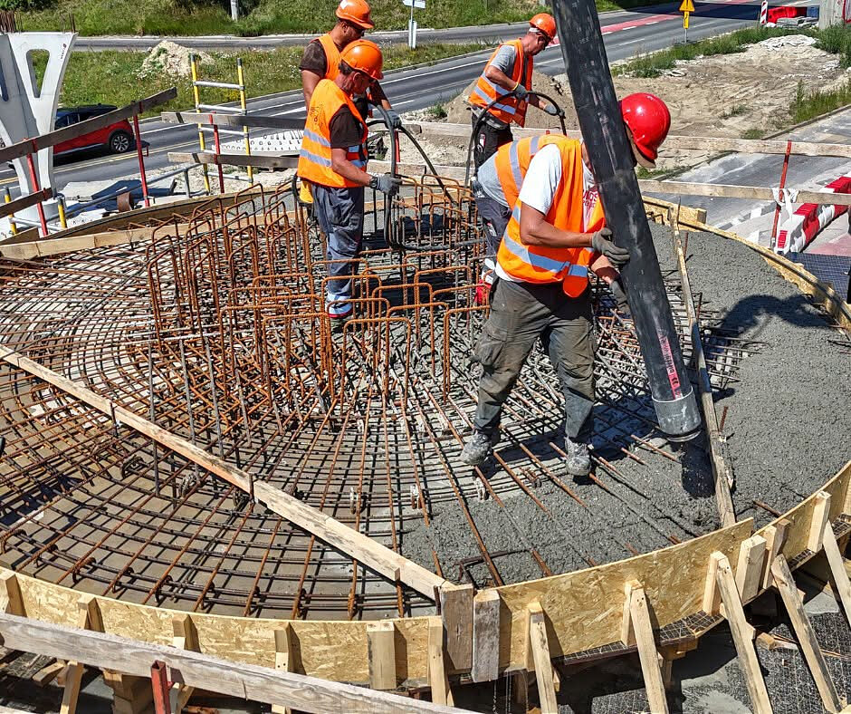 Zdjęcie przedstawia zbliżenie na robotników wylewających płynny beton z rury pompy bezpośrednio na gęstą strukturę stalowego zbrojenia. Ekipa w kaskach i kamizelkach odblaskowych pracuje wewnątrz drewnianego szalunku, który nadaje całości okrągły kształt.