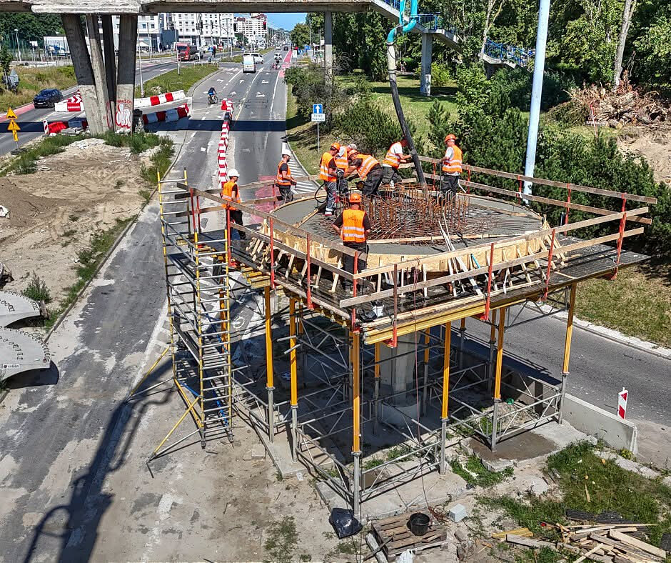 Zdjęcie przedstawia robotników pracujących na podwyższonej platformie roboczej opartej na rusztowaniach, gdzie betonowany jest okrągły element konstrukcyjny. Całość prac odbywa się bezpośrednio nad pasem wyłączonej z ruchu jezdni, w sąsiedztwie czynnego wiaduktu i infrastruktury miejskiej.