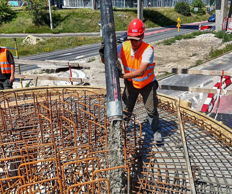 Zdjęcie przedstawia pracownika w czerwonym kasku i pomarańczowej kamizelce, który trzyma grubą rurę i precyzyjnie wylewa płynny beton. Masa betonowa trafia bezpośrednio na gęsto ułożone, stalowe pręty zbrojeniowe tworzące szkielet budowanej konstrukcji.
