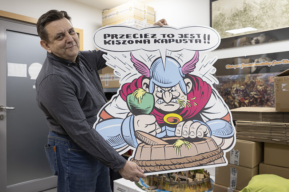 Mężczyzna w ciemnej koszuli trzyma dużą, komiksową wycinankę przedstawiającą wściekłego Gala w rogatym hełmie. Postać na planszy uderza pięścią w drewnianą beczkę, a nad nią widnieje komiksowy dymek z napisem „PRZECIEŻ TO JEST KISZONA KAPUSTA!!”.