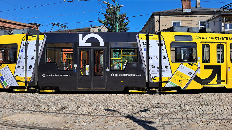 Tramwaj Czystego Miasta Gdańsk na tle zajezdni. Jest żółto-czarny i ma białe napisy