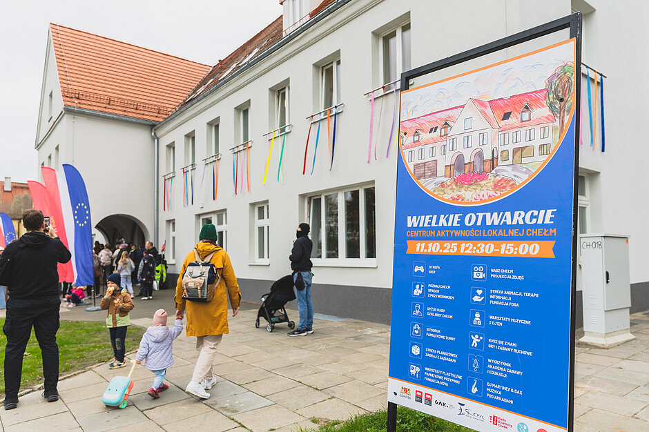 Uroczyste otwarcie Centrum Aktywności Lokalnej na Chełmie, ludzie podchodzą do budynku udekorowanego wstęgami. widoczna tablica zapraszająca na otwarcie