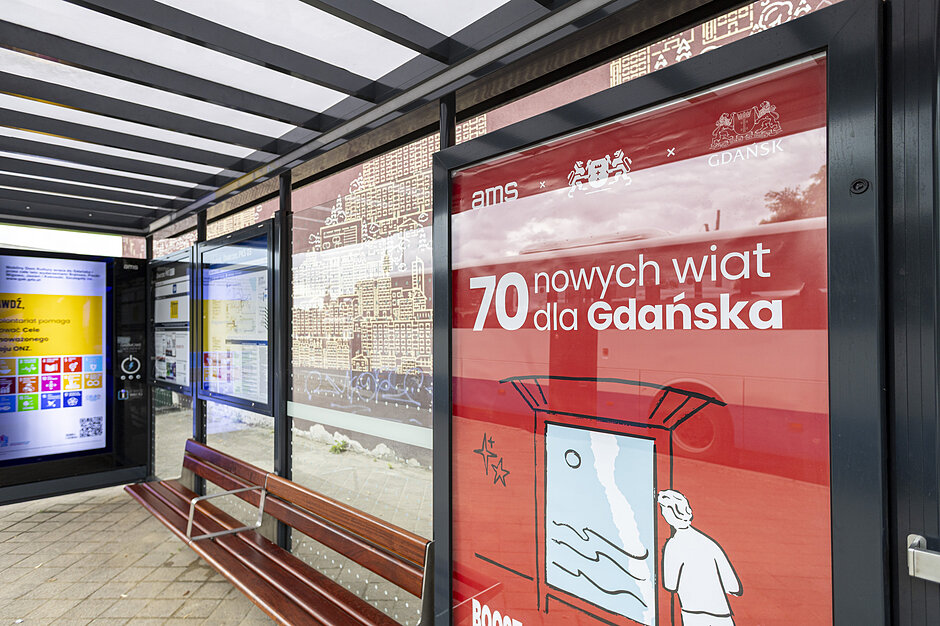 Zdjęcie przedstawia nowoczesną wiatę przystankową w Gdańsku z drewnianą ławką i szklanymi ścianami. Na pierwszym planie widoczny jest duży, czerwony plakat informujący o realizacji projektu postawienia 70 nowych wiat w mieście.