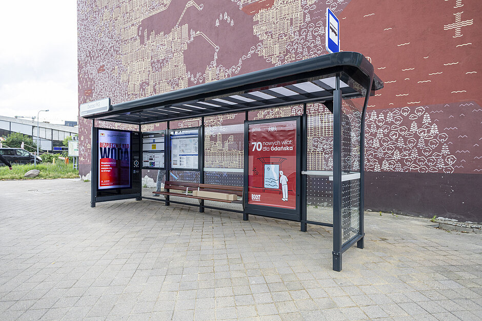 Zdjęcie przedstawia nowoczesną wiatę autobusową ustawioną na tle dużej ściany budynku pokrytej brązowym muralem. Konstrukcja przystanku jest wyposażona w podświetlane gabloty reklamowe, rozkład jazdy oraz drewnianą ławkę dla pasażerów.