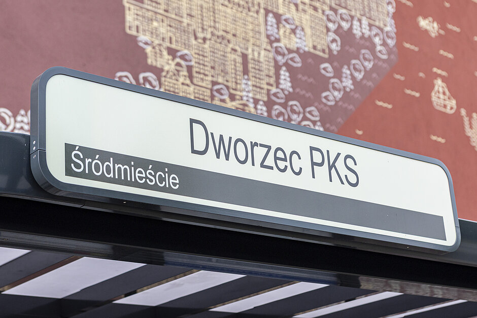 Zdjęcie przedstawia białą tablicę informacyjną umieszczoną na konstrukcji wiaty przystankowej. Widnieją na niej czarne napisy wskazujące nazwę przystanku „Dworzec PKS” oraz dzielnicę „Śródmieście” na ciemniejszym pasku.