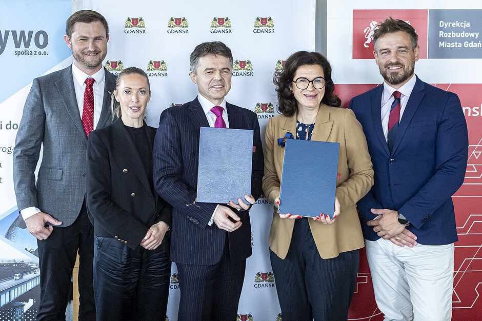Zdjęcie przedstawia grupę pięciu elegancko ubranych osób, które stoją na tle ścianki z logo miasta Gdańsk oraz Dyrekcji Rozbudowy Miasta Gdańska. Dwie osoby znajdujące się w centrum kadru trzymają w dłoniach oficjalne, ciemnoniebieskie teczki, co sugeruje moment podpisania ważnej umowy lub porozumienia.