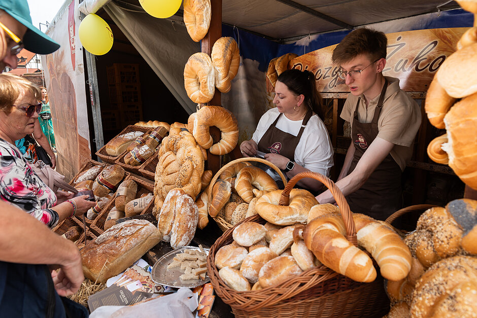 Stoisko targowe z różnorodnymi wypiekami na wystawie. Na stole znajduje się kilka koszy chlebów i ciastek, w tym bajgle, croissanty i rolki chlebowe. Stół umieszczony jest pod niebieskim namiotem z żółtymi balonami wiszącymi z sufitu. Po lewej stronie stoi kobieta w zielonym kapeluszu i okularach przeciwsłonecznych, która wydaje się patrzeć na chleb. W środku stoją za stołem dwie kobiety w fartuchach, jedna z nich trzyma kosz świeżo upieczony chleb, a druga patrzy na niego. Mężczyzna w beżowym fartużu stoi obok koszyka, obserwując wybór