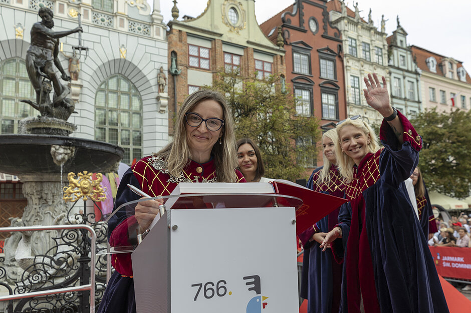 Zdjęcie przedstawia oficjalne otwarcie 766. Jarmarku św. Dominika w Gdańsku, z przewodniczącą rady miasta gdańska Agnieszką Owczarczak stojącą przy mównicy w historycznym stroju. W tle widoczna jest słynna Fontanna Neptuna oraz zabytkowe kamienice Długiego Targu, a obok mównicy stoją inne kobiety w tradycyjnych szatach.