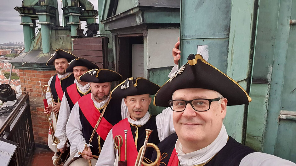 Zdjęcie przedstawia pięciu mężczyzn w historycznych strojach z trójrogimi kapeluszami, którzy robią sobie pamiątkowe selfie. Grupa znajduje się na zewnętrznym tarasie wieży, w otoczeniu ceglanych elementów architektury oraz zielonkawych, miedzianych dachów.