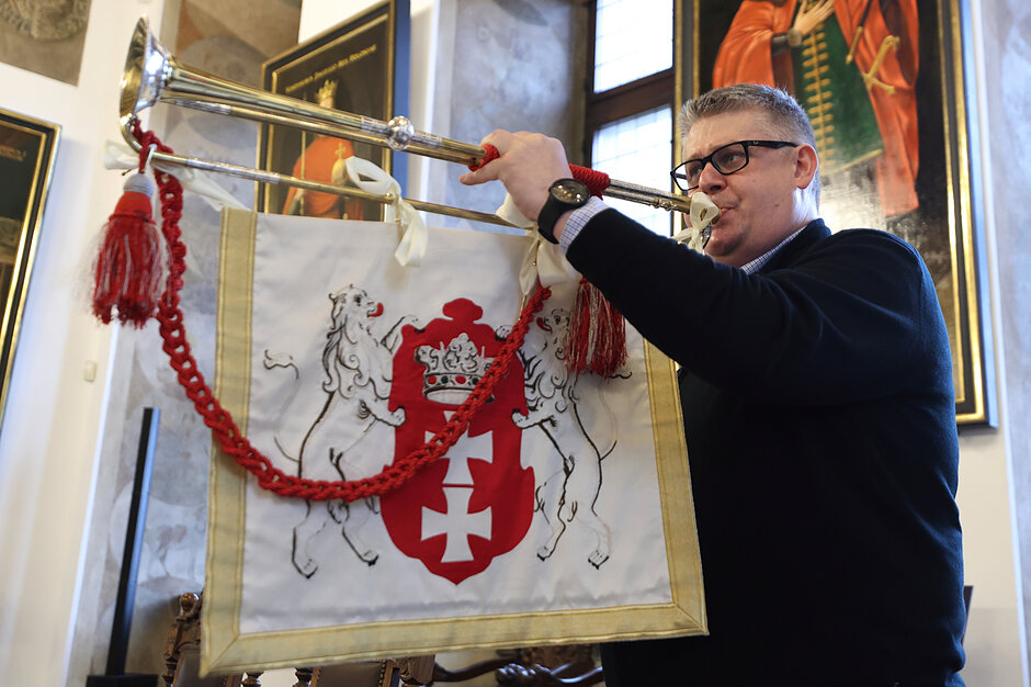 Zdjęcie przedstawia mężczyznę w okularach, który gra na długiej, ozdobnej trąbce wewnątrz zabytkowego pomieszczenia z portretami na ścianach. Do instrumentu przymocowany jest biały proporzec z wyszytym czerwonym herbem Gdańska, przedstawiającym dwa lwy podtrzymujące tarczę z dwoma krzyżami i koroną.