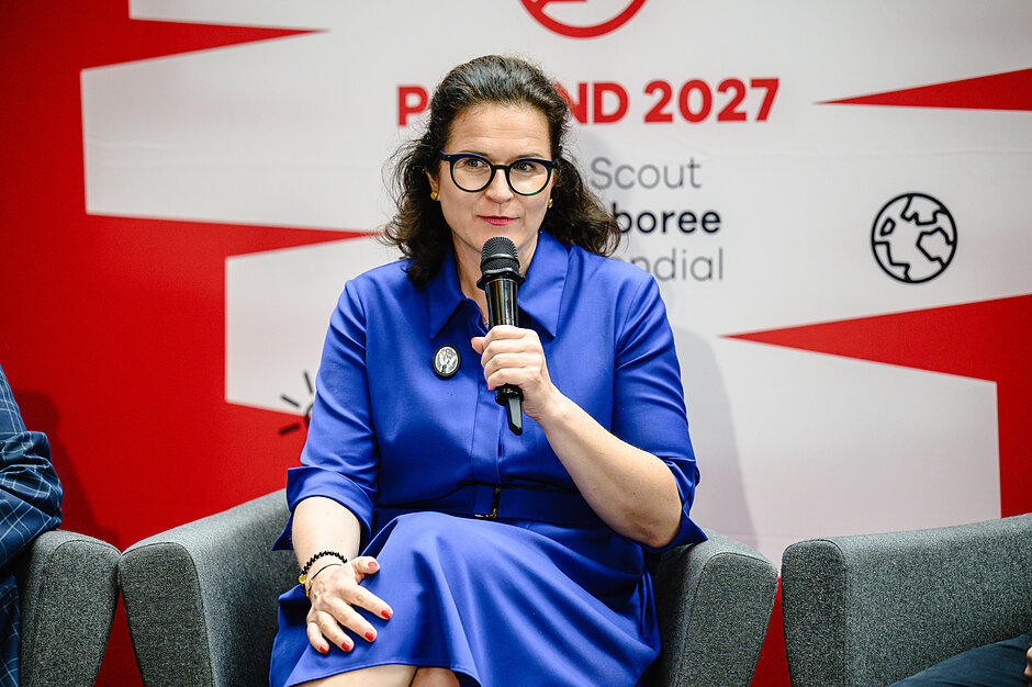 Zdjęcie przedstawia kobietę w okularach i niebieskiej sukience, która siedzi w fotelu i przemawia do trzymanego w dłoni mikrofonu. W tle widoczna jest biało-czerwona ścianka z napisami dotyczącymi Światowego Jamboree Skautowego „Poland 2027”.