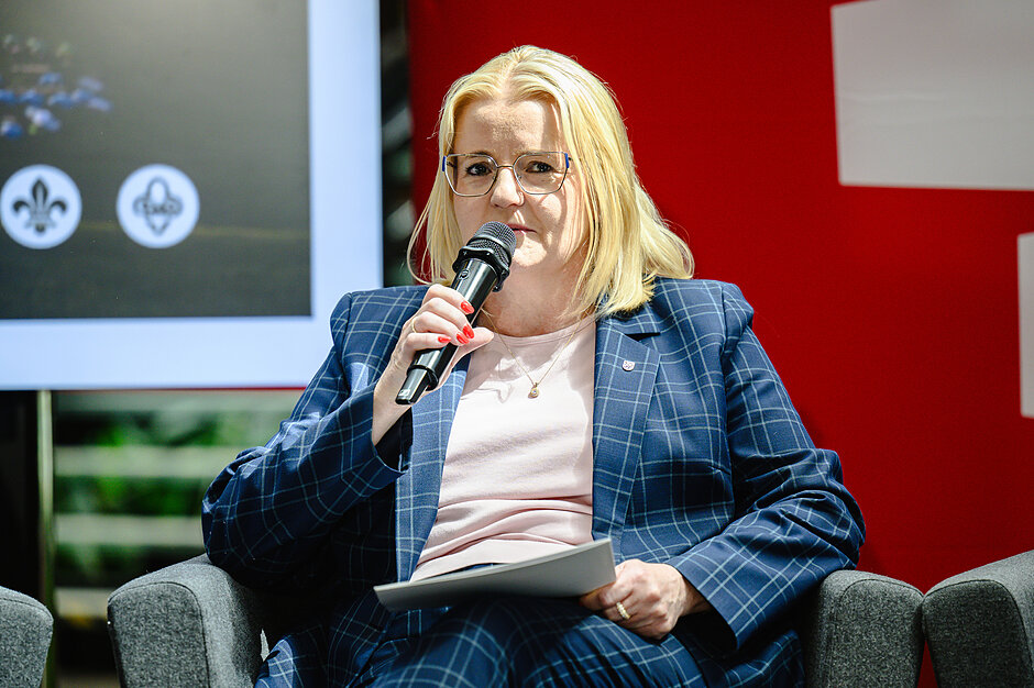 Zdjęcie przedstawia jasnowłosą kobietę w okularach i ciemnym garniturze w kratę, która przemawia do mikrofonu, trzymając w drugiej ręce kartkę papieru. Siedzi ona w szarym fotelu, a w tle po lewej stronie widoczny jest ekran z harcerskimi symbolami lilijki.