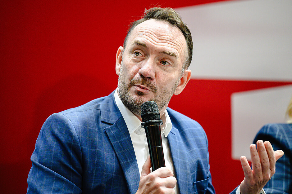 Zdjęcie przedstawia brodatego mężczyznę w niebieskiej marynarce w kratę i białej koszuli, który przemawia do trzymanego w dłoni mikrofonu. Patrzy on w bok z lekko uniesionymi brwiami, żywo gestykulując lewą ręką na tle charakterystycznej, czerwono-białej ścianki.