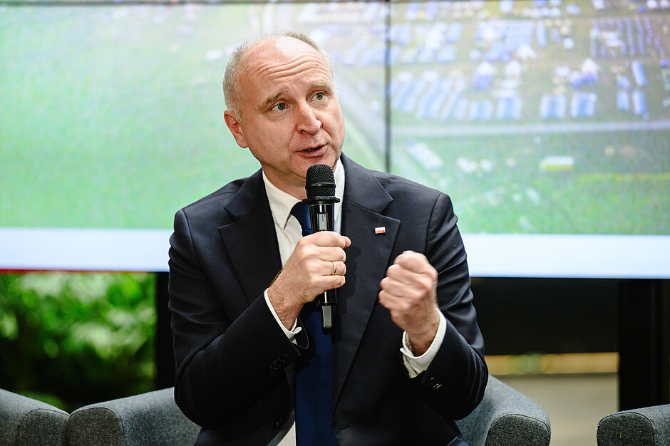 Zdjęcie przedstawia elegancko ubranego mężczyznę w garniturze i krawacie, który przemawia do mikrofonu, zaciskając lewą dłoń w pięść. Siedzi on w szarym fotelu na tle dużego ekranu, na którym wyświetla się rozmyty widok z lotu ptaka, prawdopodobnie przedstawiający teren zlotu.