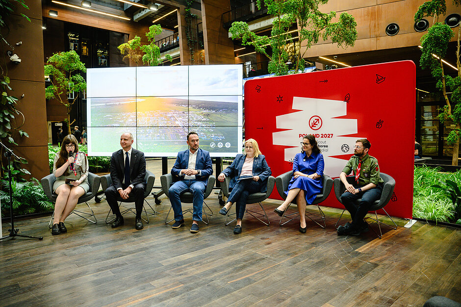Zdjęcie przedstawia sześciu uczestników konferencji prasowej siedzących w fotelach na tle dużego ekranu multimedialnego oraz czerwonej ścianki promocyjnej „Poland 2027”. Grupa, składająca się z przedstawicieli władz oraz organizacji harcerskich, dyskutuje w nowoczesnym, pełnym zieleni wnętrzu budynku.