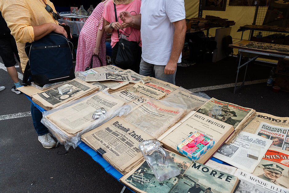 Stół pokryty niebieskim obrusem. Na stole znajduje się kilka starych gazet o różnych rozmiarach i kolorach. Gazety są rozmieszczone w sposób rozproszony, z niektórymi nakładającymi się na siebie nagłówkami i obrazami, podczas gdy inne mają tytuły takie jak Morze  i  Wielki Gatsby . Na wystawie znajdują się również książki i inne przedmioty. W tle widać dwie osoby stojące przy stołach, jedna w żółtej koszuli i niosąca czarny plecak.