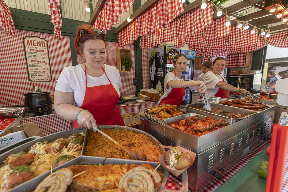 Grupa trzech kobiet w czerwonych fartuchach i białych koszulkach stojących za stołem bufetowym w restauracji. Stół jest przykryty czerwonym obrusem i na nim kilka tacy z artykułami spożywczymi. Po lewej stronie duża taca ze spaghetti, klopsami i innymi potrawami, a po prawej stronie dwie kobiety serwują sobie posiłki. Oboje trzymają tongi i wydają się być w trakcie przygotowywania potraw. W tle możemy zobaczyć deskę menu i licznik z różnymi potrawami i przyborami.