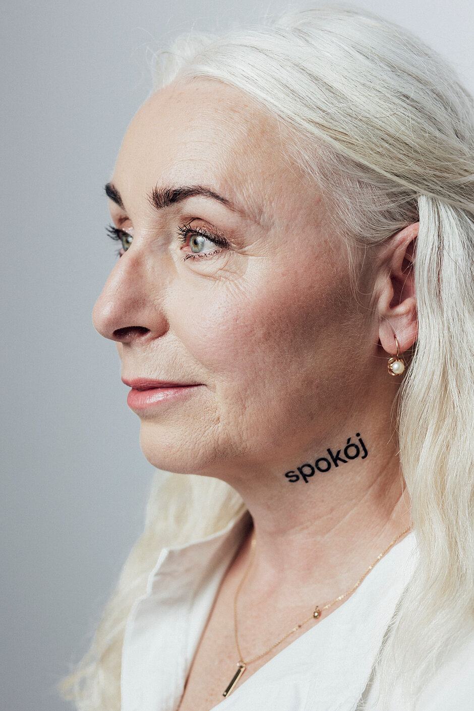 Kobieta z siwymi włosami patrzy w prawą stronę. Ma na szyi napis spokój 