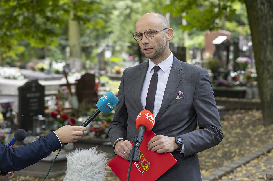 na zdjęciu mężczyzna w garniturze i okularach, mówi do mikrofonów dziennikarskich