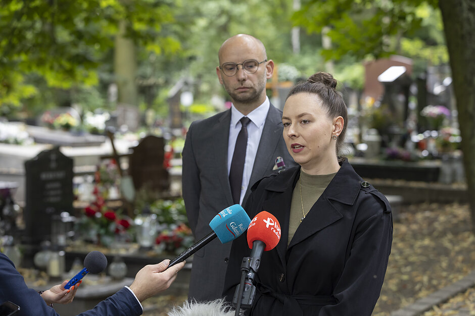na zdjęciu kobieta w garniturze mówi do dziennikarskich mikrofonów, za nią stoi mężczyzna