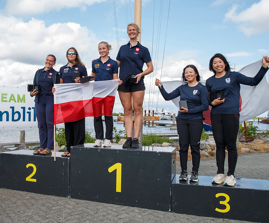 Zdjęcie przedstawia ceremonię dekoracji zawodniczek na podium, na którego najwyższych stopniach stoją reprezentantki Polski z narodową flagą. Na trzecim miejscu plasują się uśmiechnięte żeglarki z Japonii, a całe wydarzenie odbywa się na świeżym powietrzu w otoczeniu mariny.