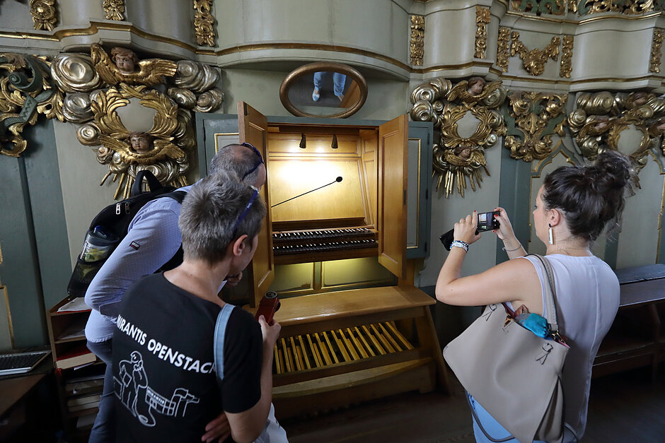 Grupa trzech osób stojących przed ozdobnym drewnianym organem w kościele. Organ jest wykonany z drewna i ma skomplikowane rzeźby na ścianach i suficie. Nad organem znajduje się duże lustro i drewniana ławka w tle. Dwójka nosi czarną koszulkę z wydrukowanymi na niej słowami OpenStack  wydają się być turystami.