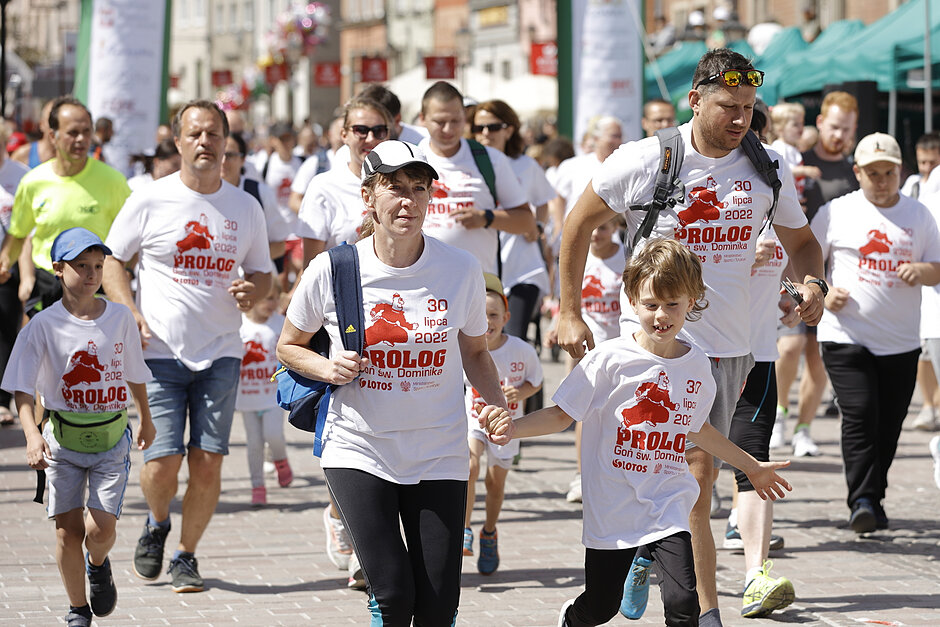 Grupa ludzi biorących udział w maratonie. Wszyscy noszą białe koszulki z wydrukowanym na nich czerwonymi literami słowem PROLOG . Ludzie chodzą po ulicy z budynkami i drzewami w tle. Niektórzy z nich noszą torby, podczas gdy inni biegną ramionami wokół siebie. W centrum jest kobieta w białej czapce i niebieskim plecaku.