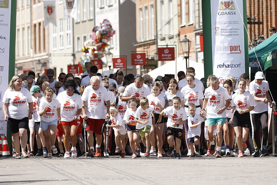 Duża grupa ludzi biorących udział w maratonie. Wszyscy mają na sobie białe koszulki z czerwonymi i zielonymi wzorami. Grupa idzie ulicą wyłożoną budynkami i banery po obu stronach są niebieskie a pogoda wydaje się słoneczna oraz ciepła.