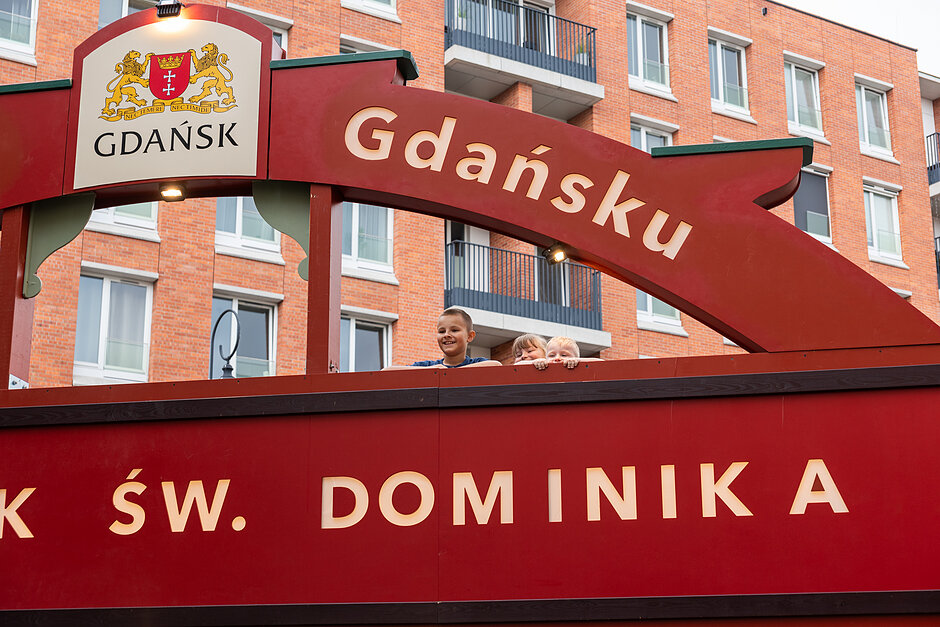 Czerwony łuk z logo Gdańska, który jest herbu znajduje się przed ceglanym budynkiem z balkonami i oknami. Pod łukiem znajdują się teksty z napisem Jarmark św. Dominika . Na pierwszym planie stoi dwóch młodych chłopców, uśmiechając się i patrząc na kamerę.
