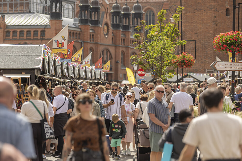 Ruchliwa ulica w Gdańsku. Wypełniona jest ludźmi w różnym wieku i pochodzenia etnicznego, wszyscy chodzącymi po chodniku. Po lewej stronie znajduje się rząd straganów sprzedających różne przedmioty, takie jak flagi, banery i inne przedmiotów dekoracyjnych. W tle możemy zobaczyć duży ceglany budynek z łukowymi oknami i wieżą zegarową.