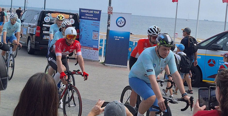 kolarze i kolarki jadą na rowerach, pięć osób. Za nimi plakat informujący o akcji transplant tour