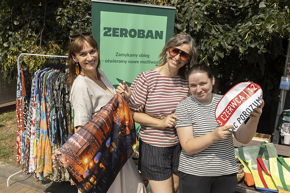 trzy kobiety pozują, pokazując torby, za nimi baner z napisem Zeroban oraz wieszak z długimi kolorowymi spódnicami