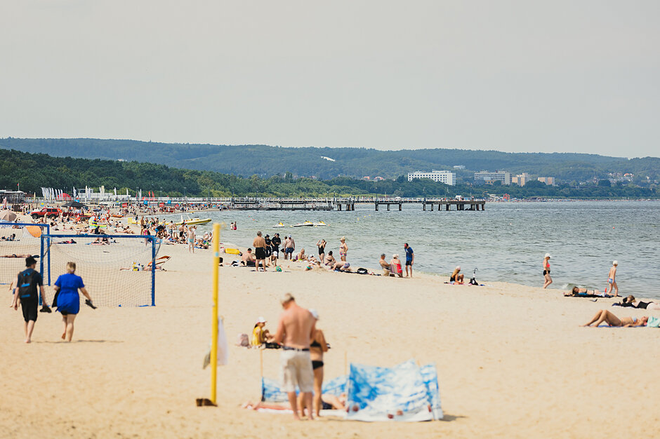 Zatłoczona plaża z wieloma ludźmi cieszącymi się słońcem i piaskiem. Plaża znajduje się w pobliżu wody i jest molo rozciągające się w odległości. W tle znajdują się wzgórza pokryte drzewami i budynkami. Po lewej stronie obrazu widzimy grupę ludzi spacerujących wzdłuż plaży, niektórzy z nich grają w siatkówkę, podczas gdy inni leżą na piasku i cieszą się sobą. Jest też kilka osób siedzących na krzesłach i parasolkach rozproszonych wokół brzegu. Niebo jest niebieskie, a pogoda wydaje się słoneczna i ciepła, sugerując, że zdjęcie zostało zrobione w słoneczny dzień.