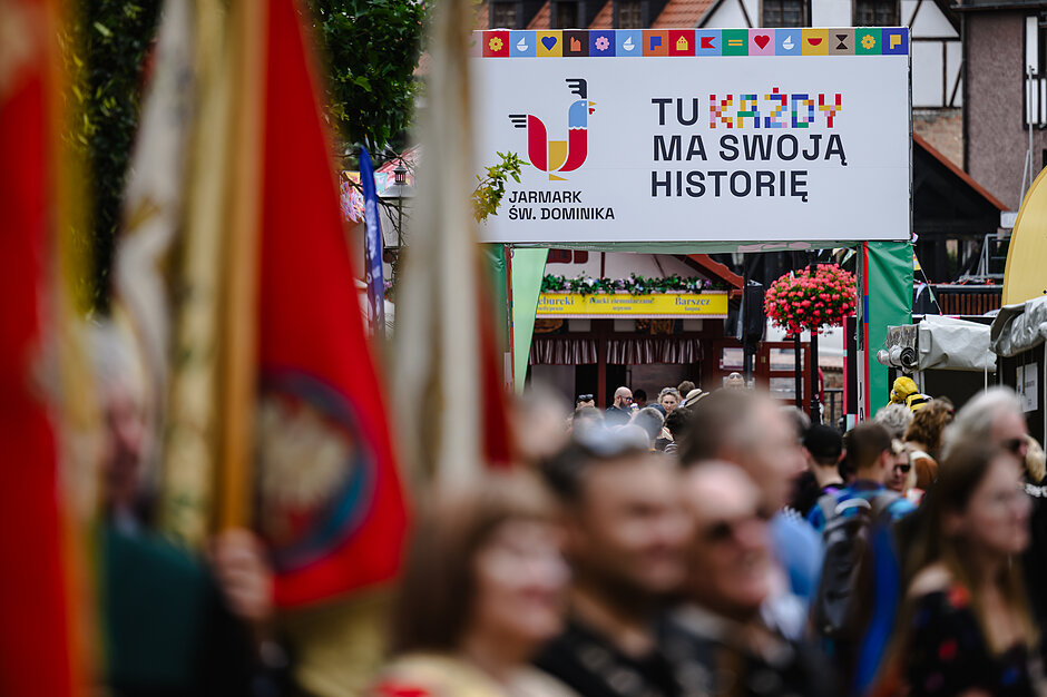 Zdjęcie przedstawia tłum ludzi zgromadzonych na tradycyjnym Jarmarku św. Dominika, nad którym góruje biały transparent z hasłem „Tu każdy ma swoją historię”. Na pierwszym planie widoczne są rozmyte, czerwono-białe polskie flagi, które nadają ujęciu uroczysty charakter.