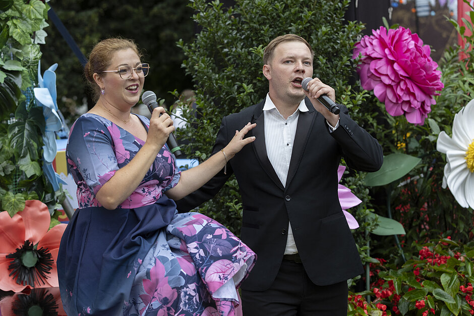 mężczyzna i kobieta stojący w ogrodzie z różnymi kolorowymi kwiatami i roślinami; kobieta po lewej stronie trzyma mikrofon i wydaje się, że mówi do niego, podczas gdy mężczyźni stoją obok niej z otwartymi ustami i rękami gestami, jakby śpiewali; oboje są ubrani w formalny strój, z kobietą w okularach, a mężczyzną w garniturach; w tle jest duży różowy kwiat dalii i biały kwiat daisy, oraz trochę zieleni oraz drzew; wydaje im się, iż wykonują piosenkę lub przedstawienie.