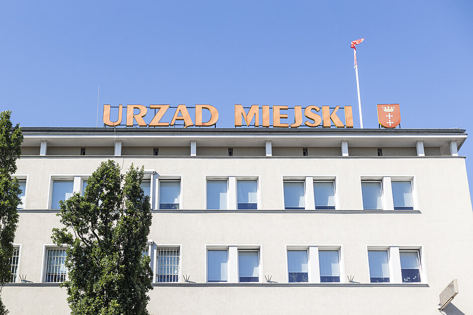Zdjęcie przedstawia jasną elewację budynku Urzędu Miejskiego w Gdańsku na tle bezchmurnego, niebieskiego nieba. Na dachu obiektu zamontowano duży, pomarańczowy napis z nazwą instytucji oraz herb miasta z flagą na maszcie.