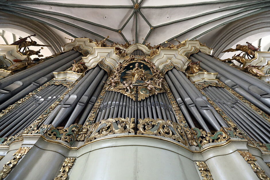 Wnętrze dużego, ozdobnego organu rury w kościele; organ składa się z wielu rzędów rur, każdy o skomplikowanych złotych i srebrnych wzorów i wzorów; nad organem znajduje się centralna postać Jezusa Chrystusa, z wyciągniętymi ramionami i aureolą nad głową; ma na sobie koronę i pałeczkę; po obu stronach figury są dwa anioły, po jednej stronie, trzymające ręce, a po drugiej na szczycie siebie; organ wydaje się być wykonany z metalu i ma wysoki sufit w kształcie krzyża; jest też kilka posągów świętych i aniołów na obu końcach; na dole tej kolumny organu ściany każdej kolumnie są pomalowane na biało, i są kolumnami podtrzymującymi strukturę.