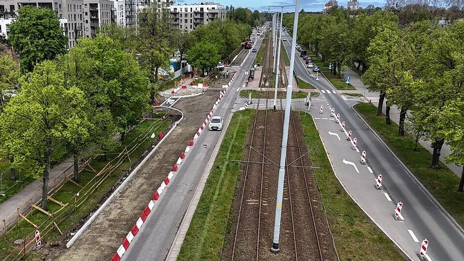 Zdjęcie z lotu ptaka na tory tramwajowe i jezdnię, wzdłuż których rosną po obu stronach drzewa. W tle bloki mieszkalne.