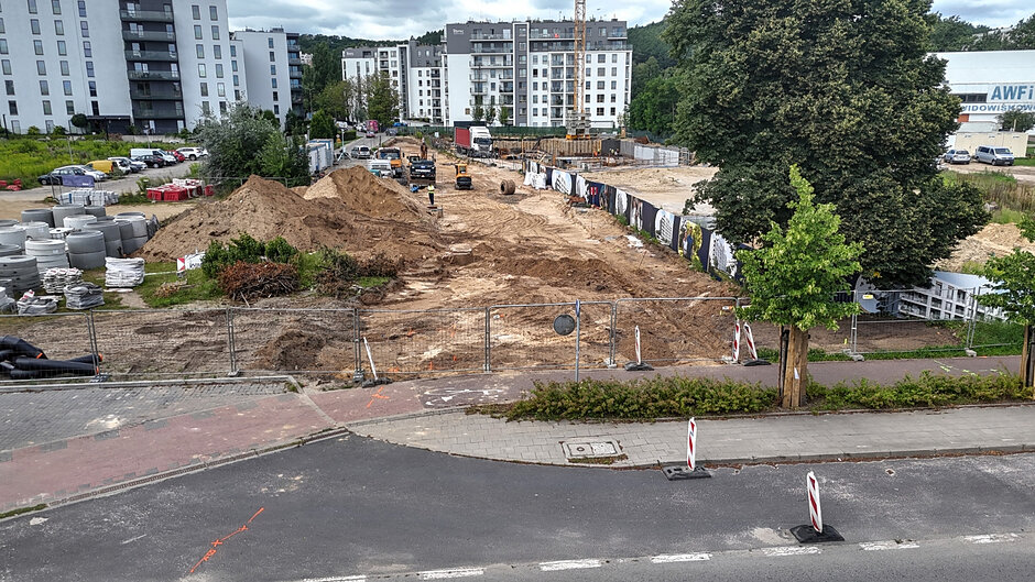rozkopany teren, widać hałdy piasku, w tle kilka bloków mieszkalnych, kilkunastopiętrowych o jasnej elewacji