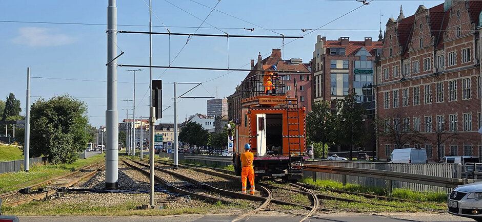na zdjęciu dwóch robotników w kombinezonach stoją na dachu pomarańczowego samochodu, na torach tramwajowych, montują sieć trakcyjną przy słupach
