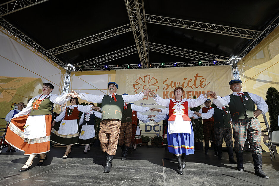 grupa ludzi wykonujących taniec na scenie. Na zdjęciu znajduje się sześć osób, wszystkie ubrane w tradycyjne bawarskie ubrania. Trzymają się za ręce i tańczą w kręgu, z wyciągniętymi przed nimi ramionami. 