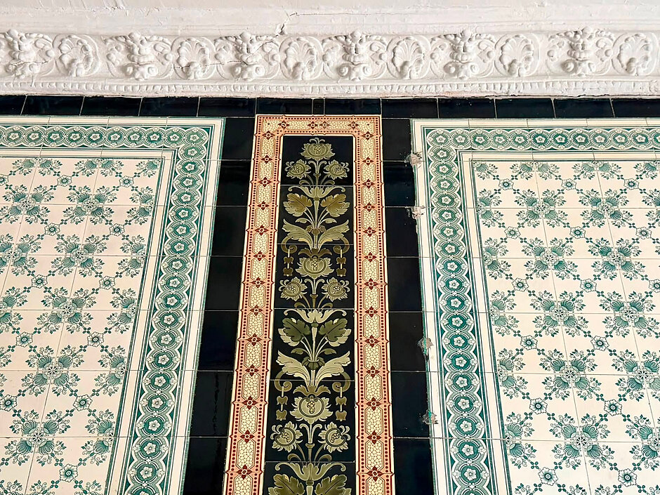 Zdjęcie przedstawia zabytkową ścianę wyłożoną ozdobnymi płytkami ceramicznymi o bogatych, roślinnych i geometrycznych wzorach w odcieniach zieleni, czerni i beżu. W górnej części kadru widoczny jest fragment białej ściany zwieńczonej dekoracyjnym, rzeźbionym gzymsem ze sztukaterią.