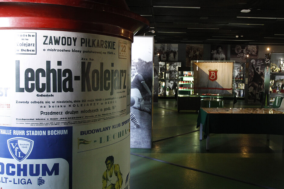duży czerwono-biały cylindryczny obiekt z etykietą. Na etykiecie znajduje się napis "Zawody Piłkarskie Lechia-Kolejarz". Pod etykietą znajdują się zdjęcia piłkarza i logo polskiej reprezentacji piłkarskiej. 