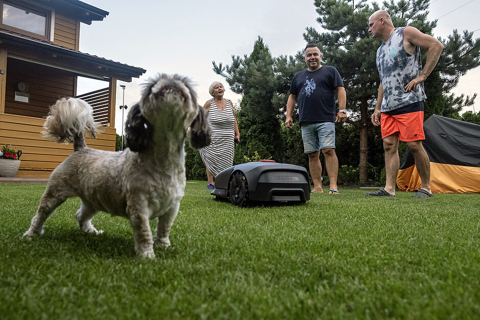 Kobieta i dwóch mężczyzn stoją w ogrodzie na działce ROD, po trawniku jeździ robot kosiarka, obo stoi pies.
