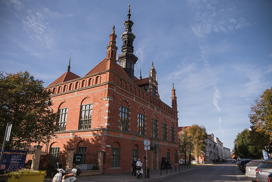 duży budynek z czerwonej cegły z dwoma wysokimi igłami na górze budynek wydaje się być kościołem lub podobną strukturą z ostrym dachem i ozdobnymi detalami znajduje się na ulicy z samochodami zaparkowanymi po prawej stronie i kilkoma osobami chodzącymi i siedzącymi na ławkach drzewa po obu stronach są niebieskie z kilkoma chmurami w tle widoczne są inne budynki.