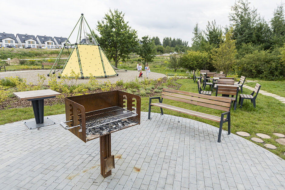 Przestrzeń rekreacji na osiedlu, na pierwszym planie grill, ławka i stolik, dalej plac zabaw i dzieci.