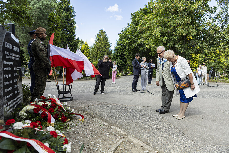 na zdjęciu dwie starsze osoby kłaniają się przed czarnym pomnikiem, przed którym leżą wiązanki kwiatów